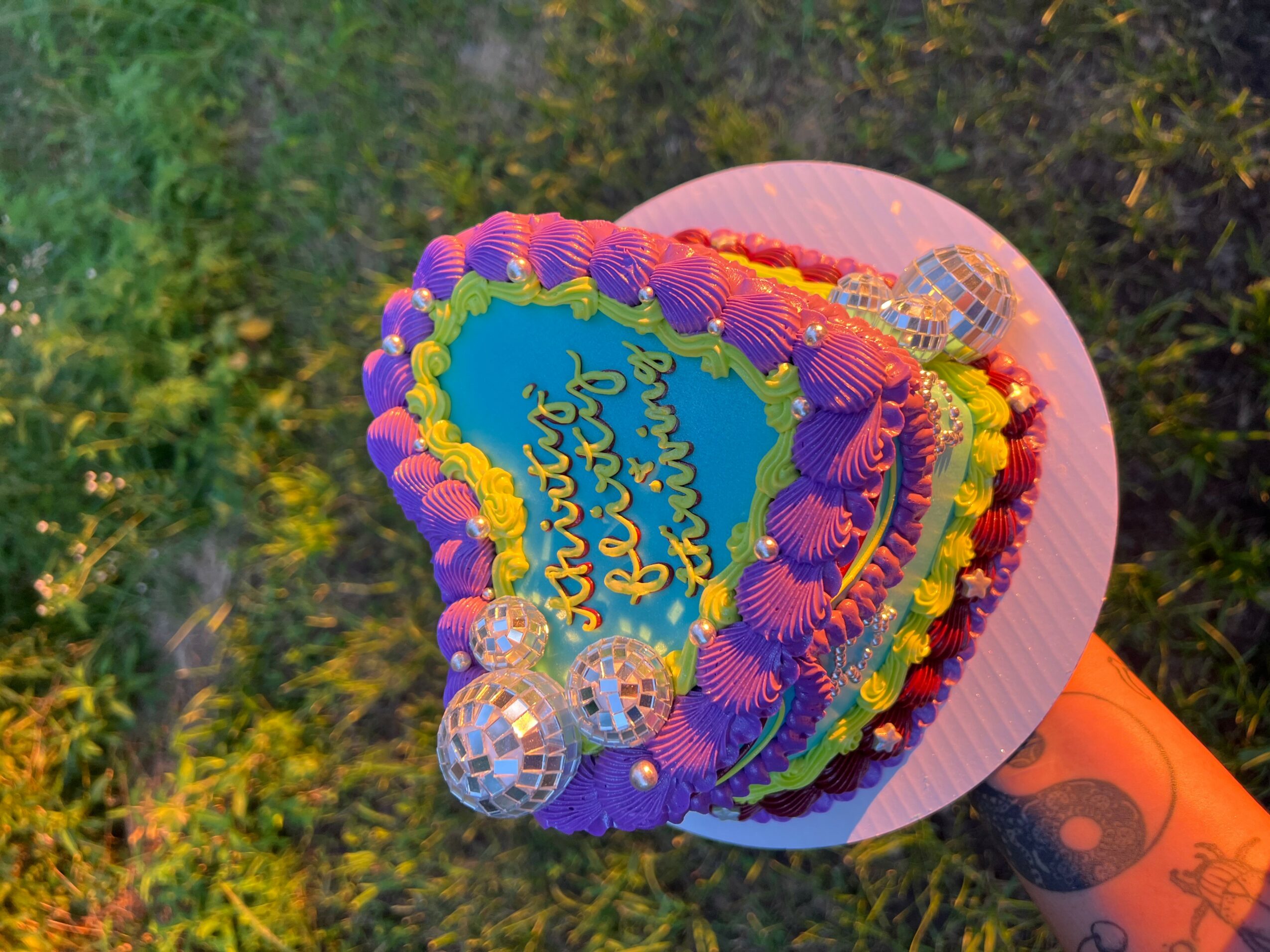 Heart-shaped cake with colorful frosting, decorations, and a message, on a pink base, outdoors with trees in background.