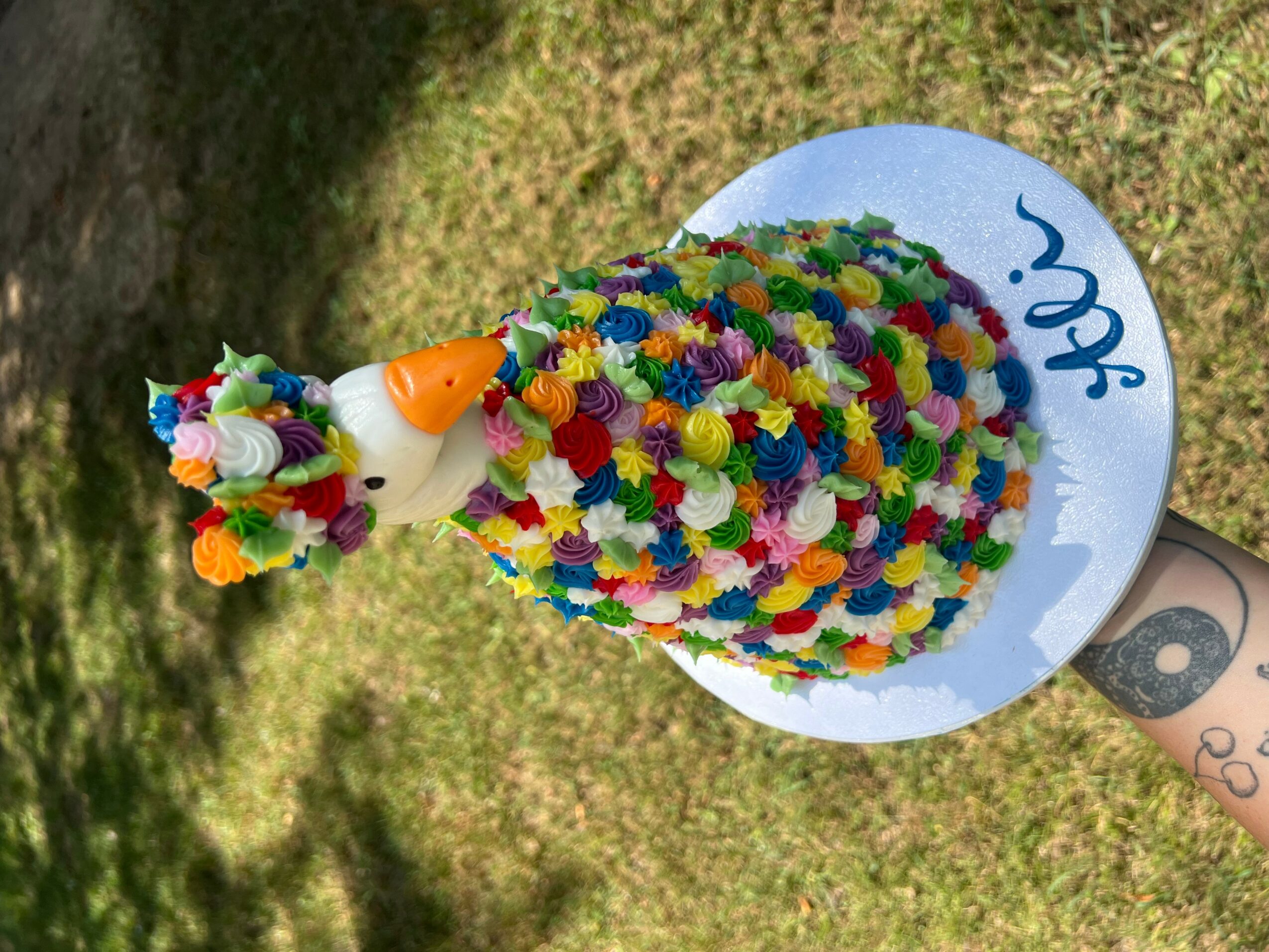 Colorful cake with a unicorn decoration on top, placed on a white plate outdoors on grass.