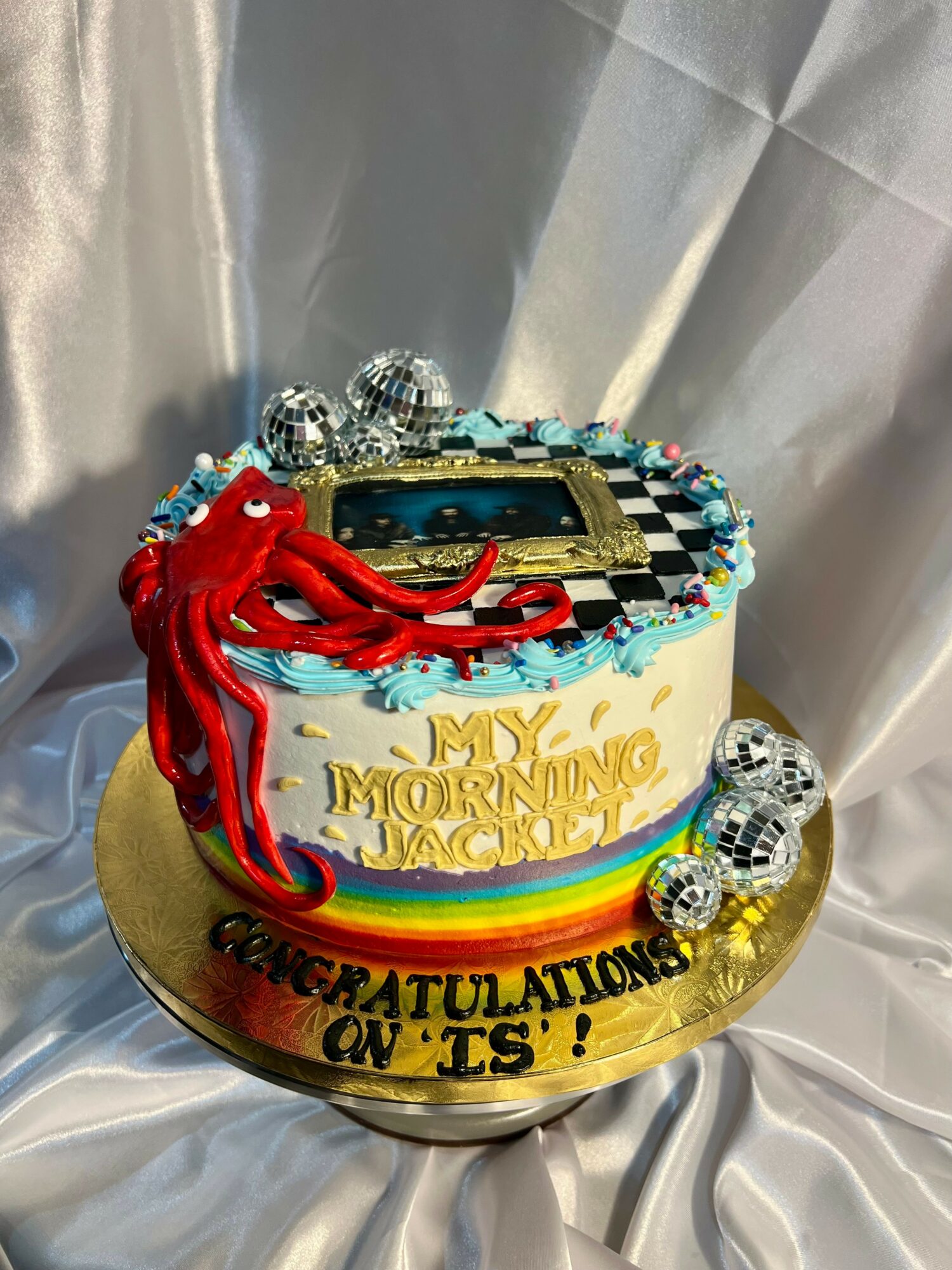 Round cake with a rainbow-colored band, decorated with a red octopus, disco balls, and a gold message, on a gold base.