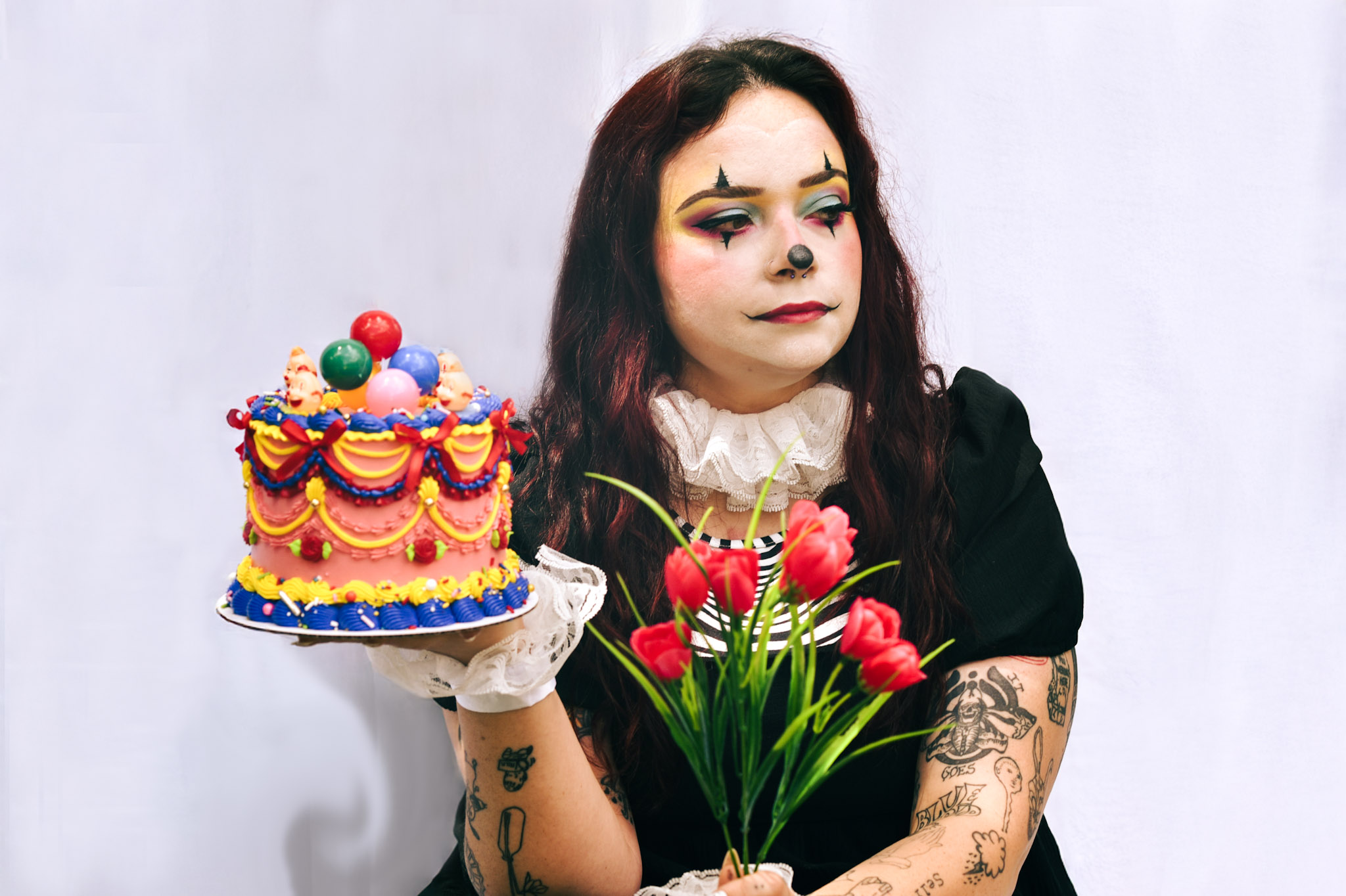 A woman with dark hair and tattoos holds a bouquet of pink tulips, with a colorful cake in the background.