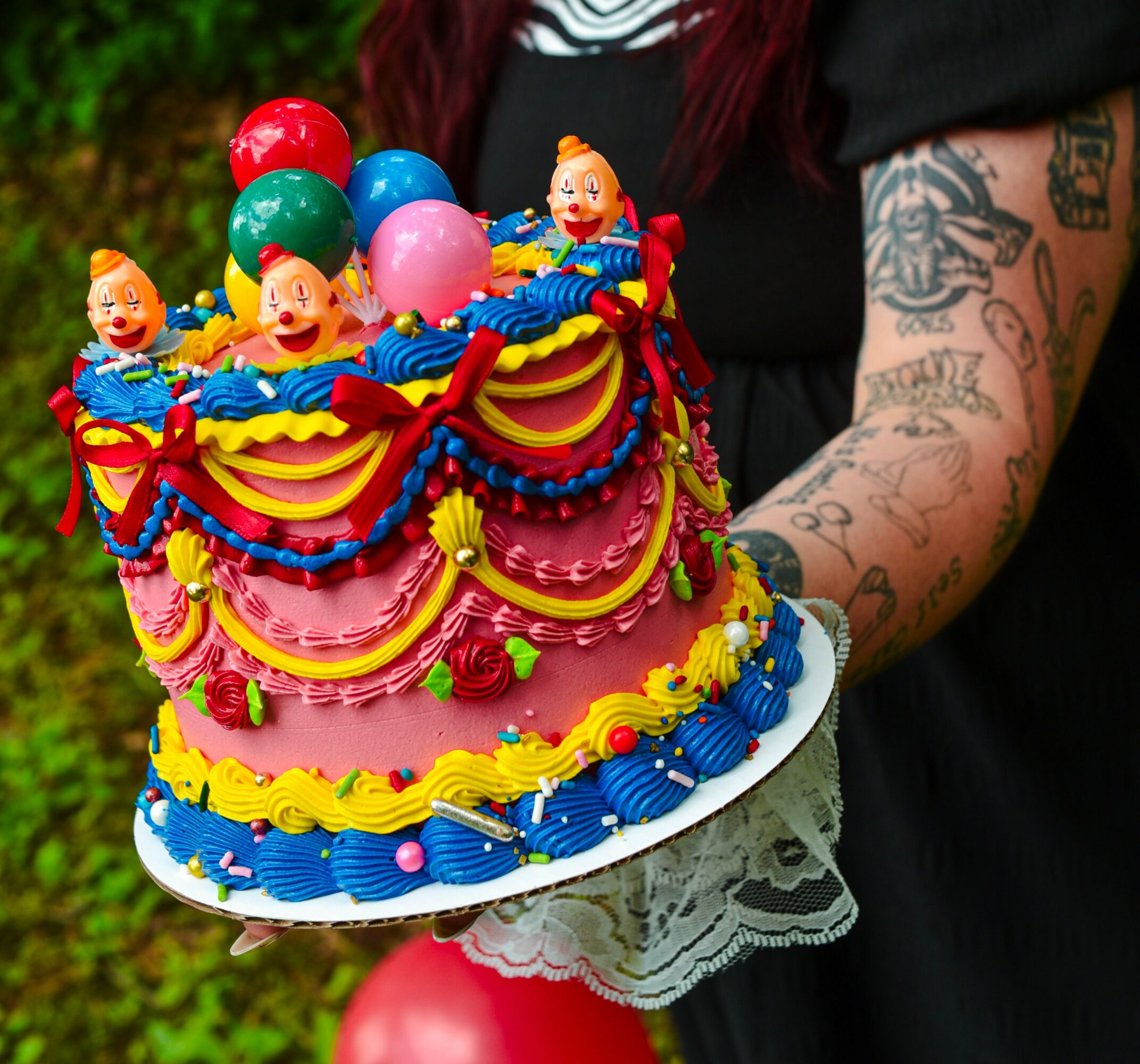 Person holding a colorful decorated birthday cake with clown figurines and balloons, outdoors, with tattoos visible on arm.