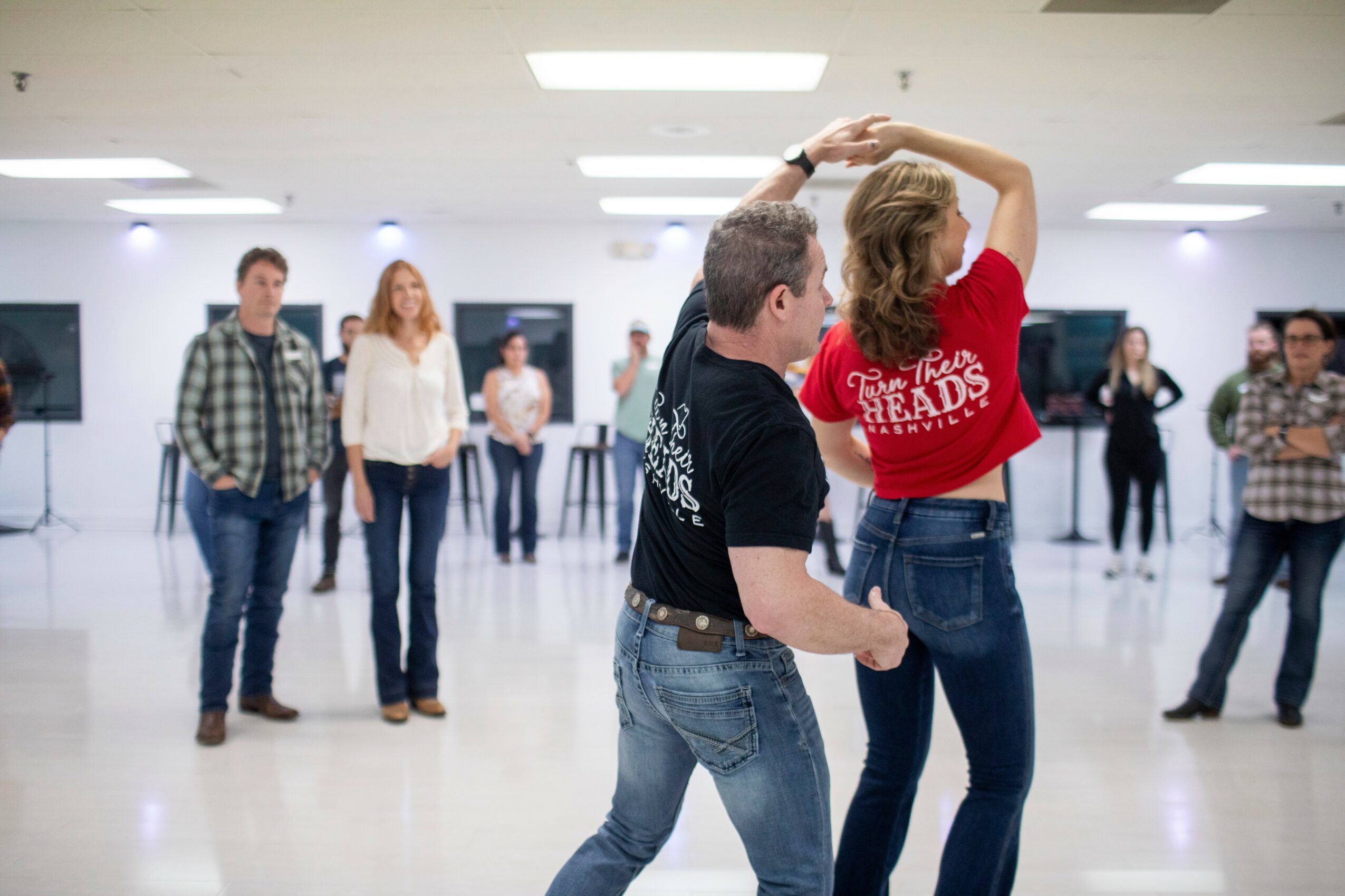 Two people dance in a spacious room with others watching, bright lighting, and a plain background.