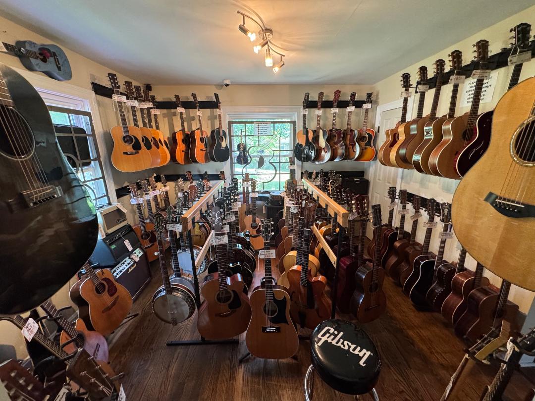 Room filled with guitars on walls and stands, window at back, ceiling light, wooden floor, music equipment visible.