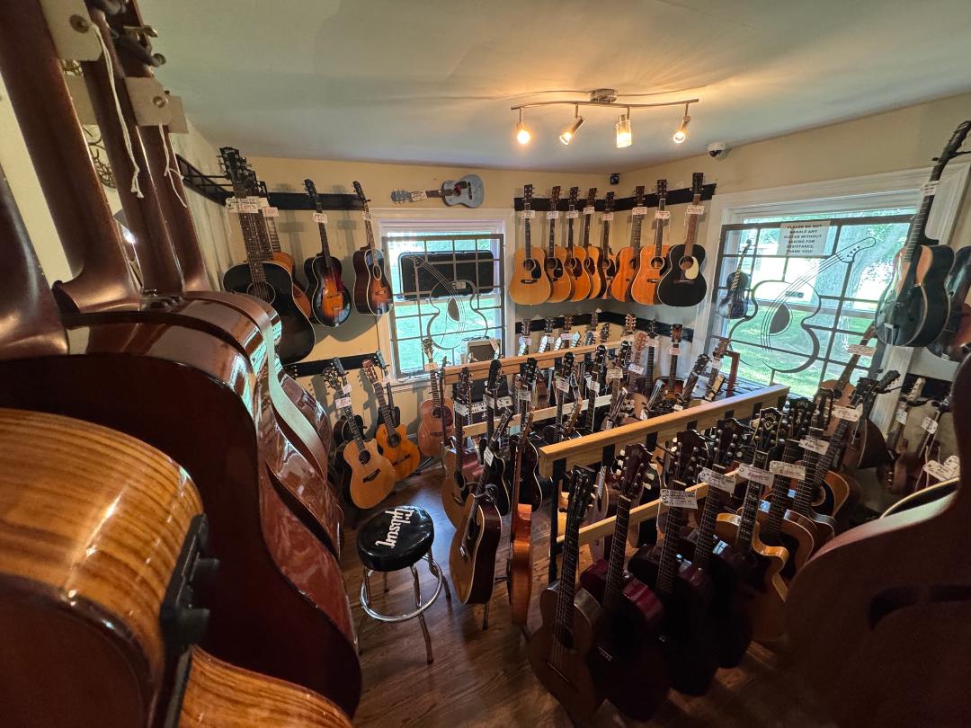 Room filled with guitars hanging on walls and standing on racks, with a window and ceiling lights, viewed from a corner.