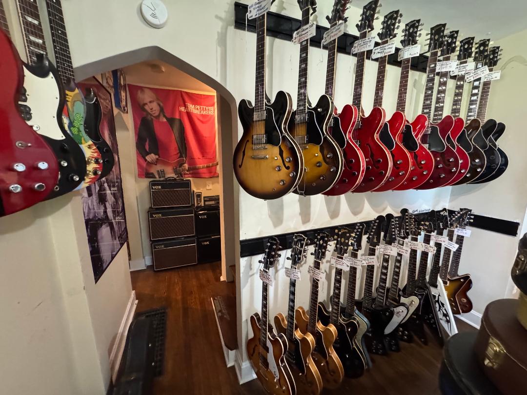 Guitars hanging on a wall in a music store, with a poster and stereo system visible in the background.