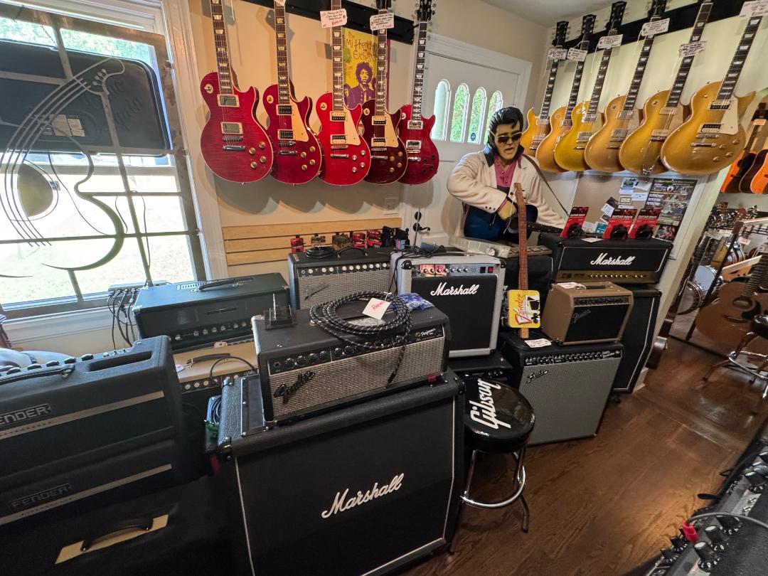 Person standing in a music store surrounded by guitar amplifiers and hanging guitars on the wall, with a window on the left.