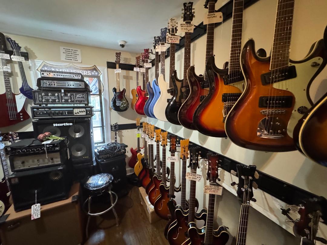 Room with guitars hanging on wall, amplifiers, and music equipment, with a window and a stool in the corner.