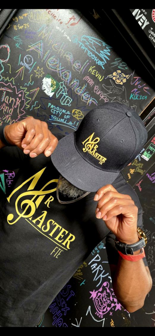 Person wearing a black cap and hoodie with yellow text, holding the cap's brim, in front of a chalkboard wall with colorful drawings.