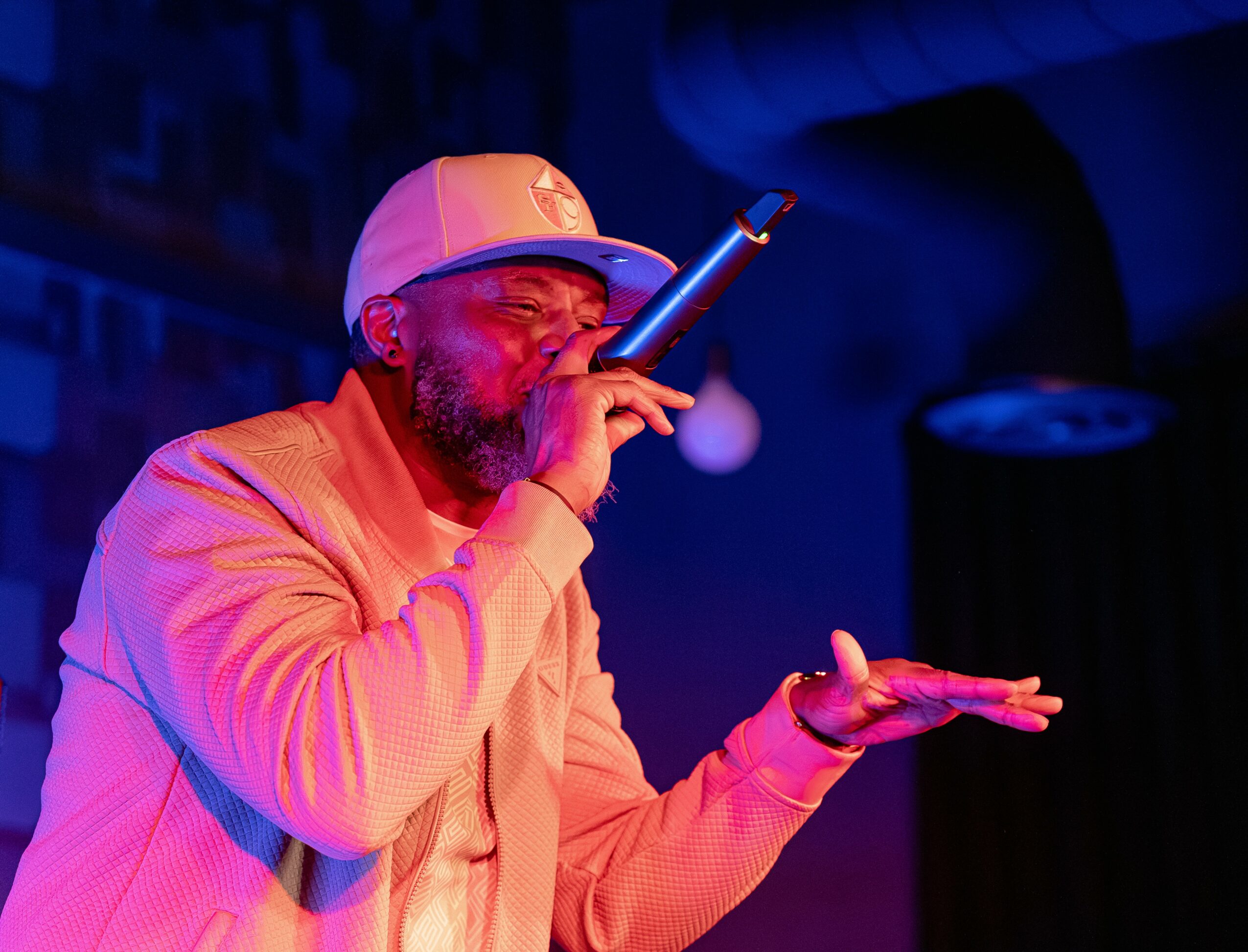 Person wearing a cap and glasses, holding a microphone and performing on stage with colorful lighting.