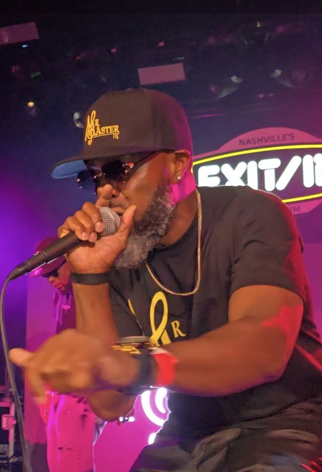 Man with beard wearing sunglasses, a cap, and a black t-shirt, holding a microphone on stage with colorful lights.