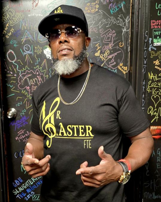 Man with sunglasses, beard, black cap, black T-shirt, gold chain, and watch, standing in front of a colorful chalkboard wall.