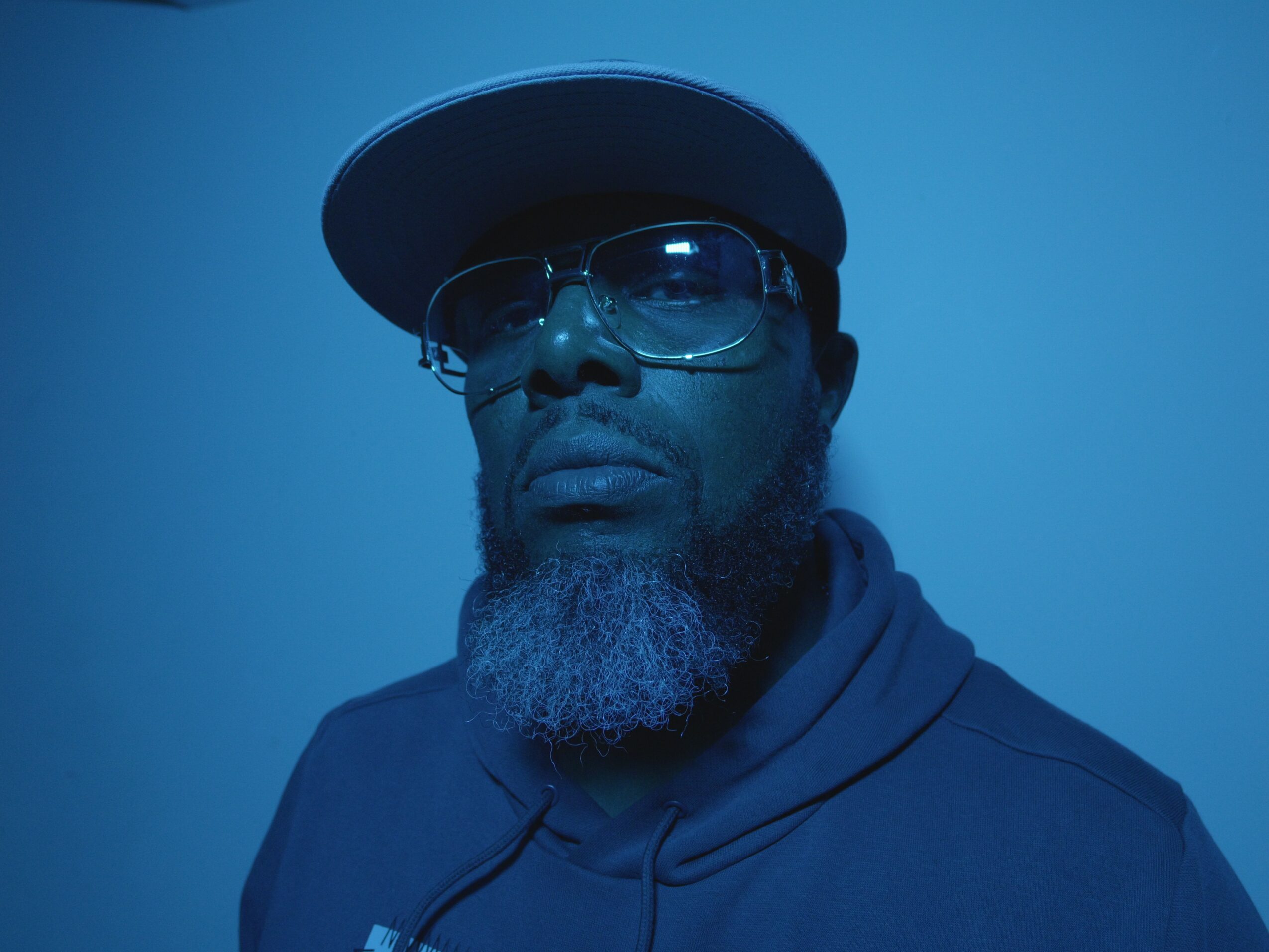 Portrait of a man with glasses, a cap, and a beard, against a blue background.