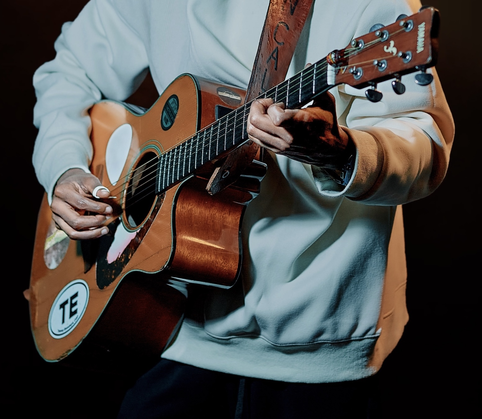 Person playing an acoustic guitar, wearing a light-colored hoodie, with a dark background.