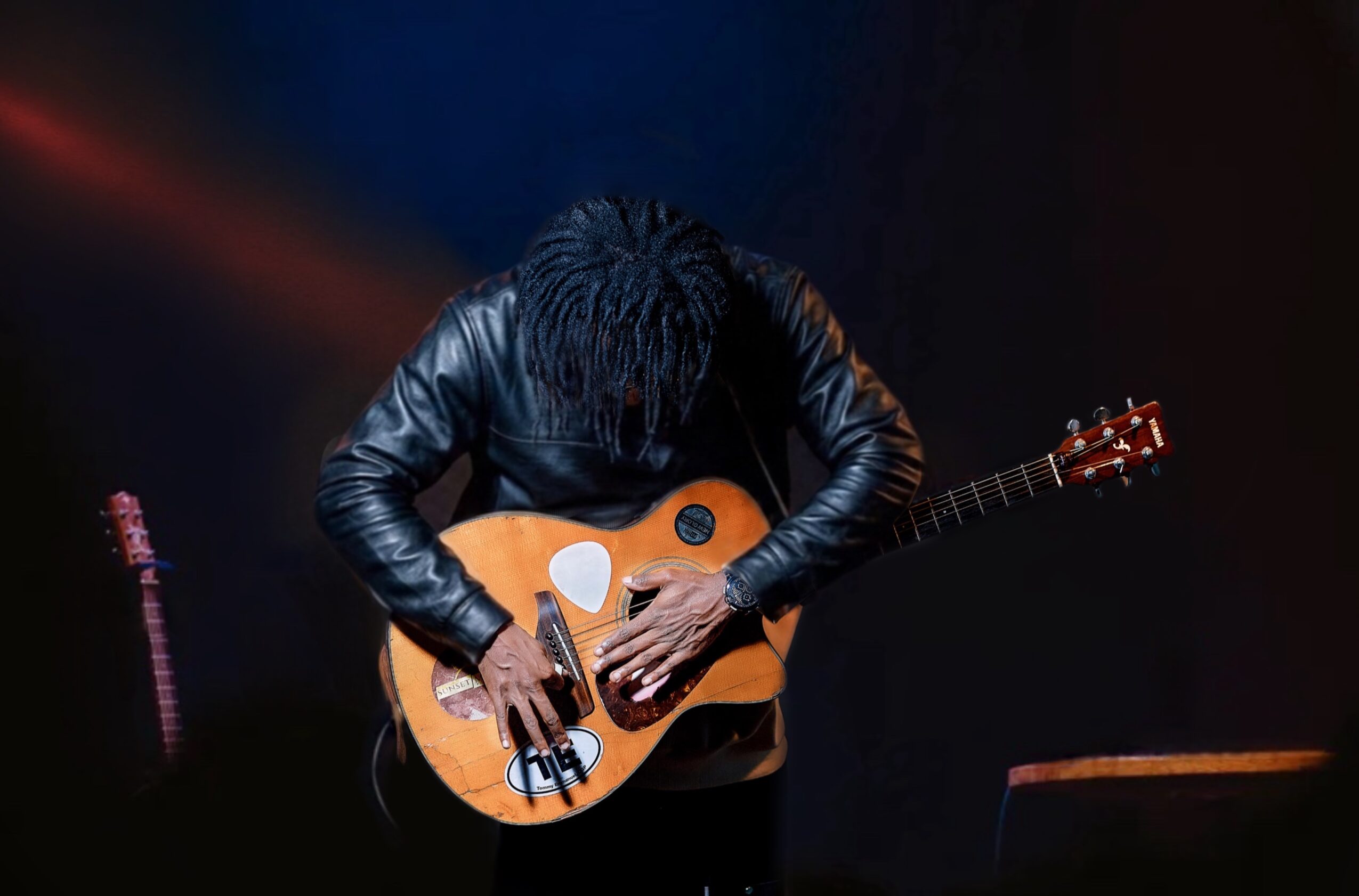 Person playing an acoustic guitar on stage, head down, dark background, wearing a leather jacket.