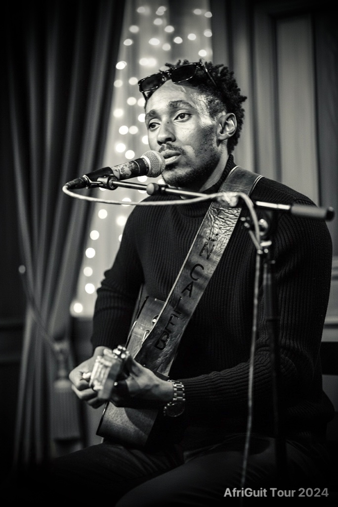 Male musician singing into a microphone while playing an acoustic guitar on stage, with curtains and lights in background.