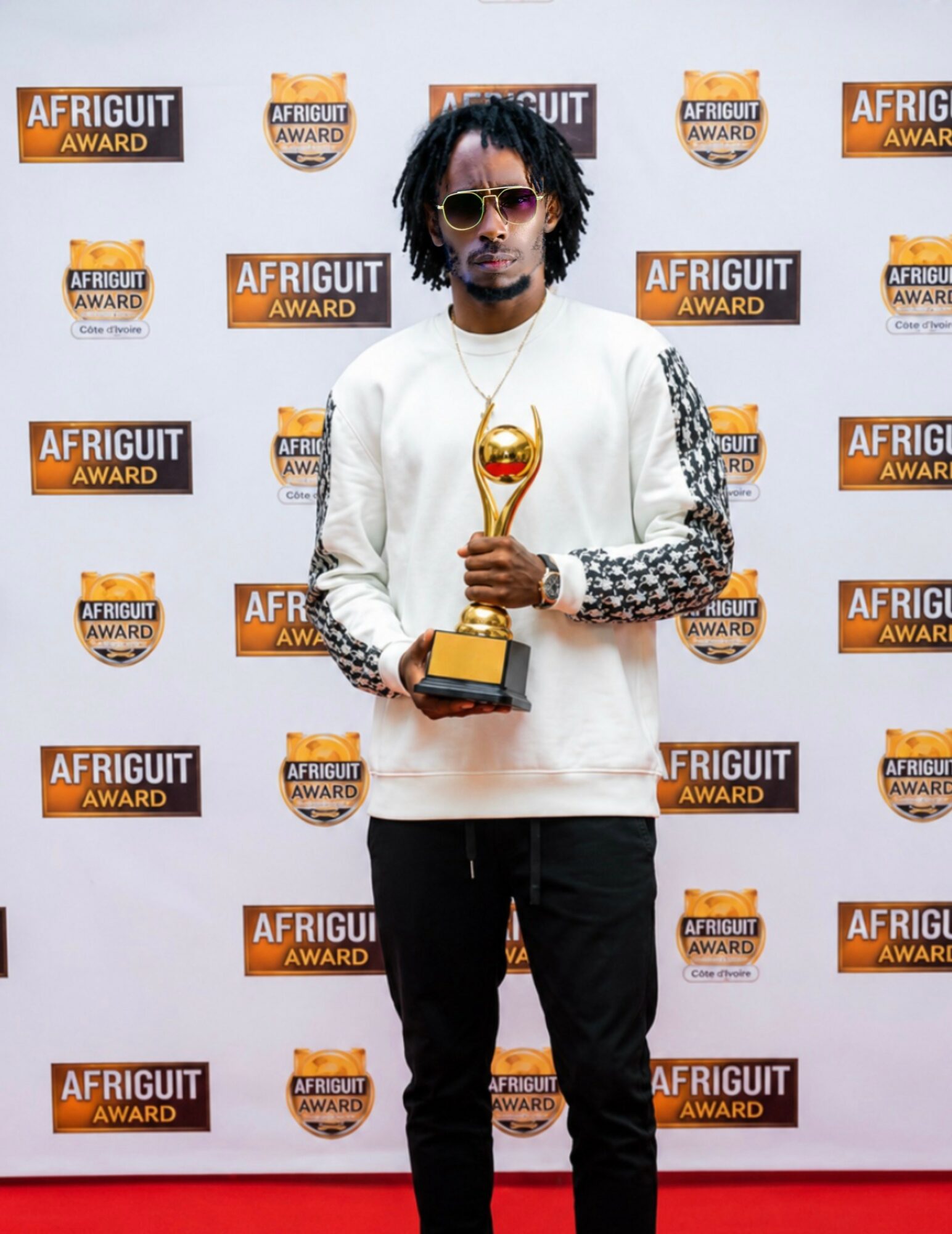 Man with sunglasses and curly hair holding a trophy in front of a backdrop with 'AFRIGUIT AWARD' logos.