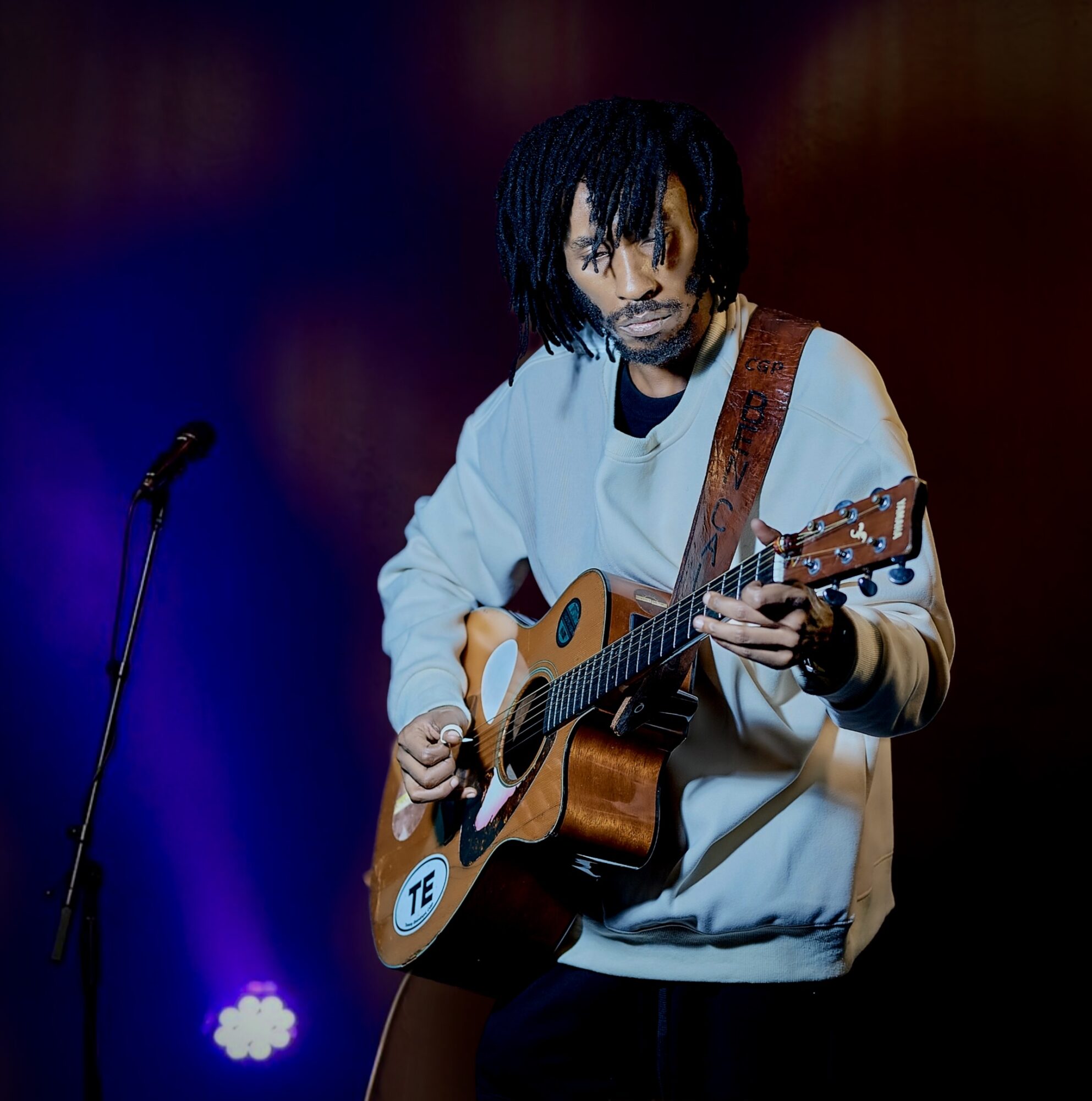 Person with dreadlocks playing an acoustic guitar on stage, with a microphone nearby, dark background, colorful lighting.