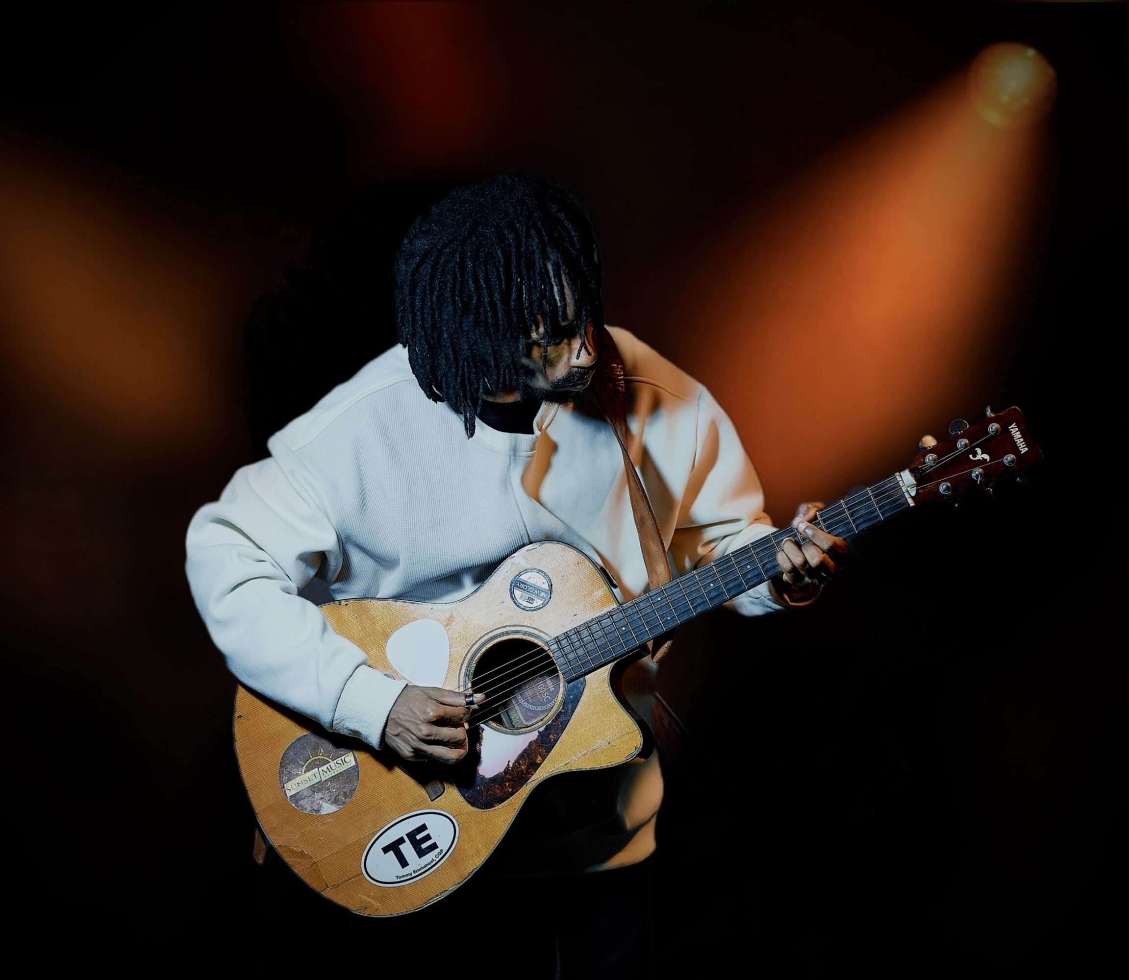 Person with dark hair playing an acoustic guitar on stage with orange lighting in background.