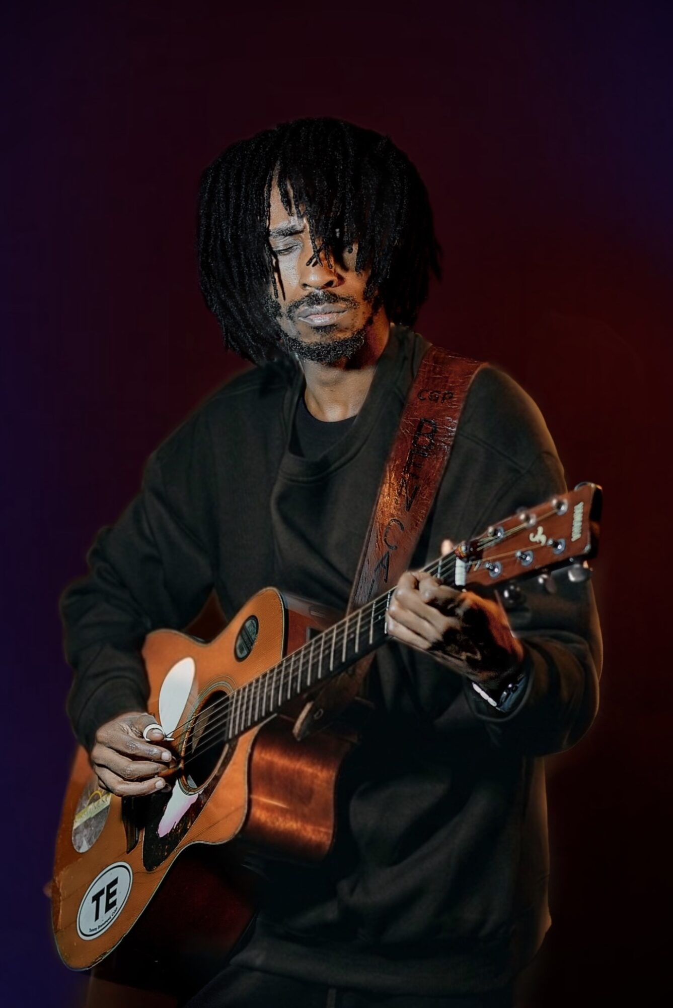 Person with dreadlocks playing an acoustic guitar, wearing a dark jacket, against a dark background.