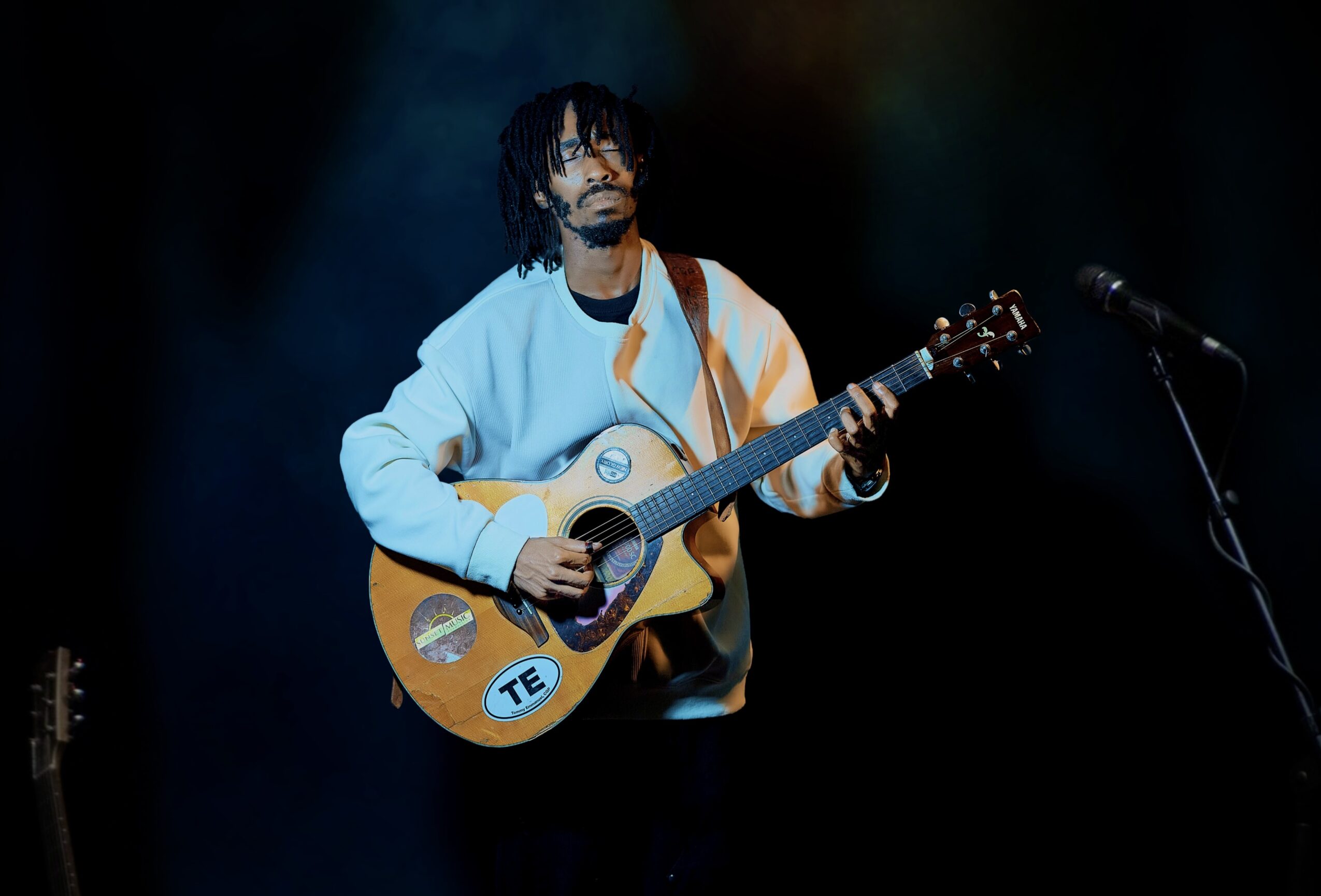 Person with dreadlocks playing an acoustic guitar on stage with a microphone nearby, dark background.