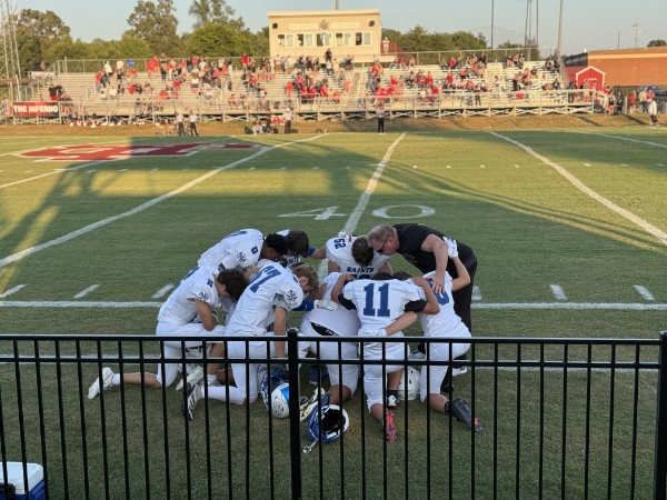 Group of football players kneeling and praying on the field, with spectators in the background.