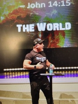 Person speaking at a podium with a water bottle, background displays text 'John 14:15' and 'THE WORLD'.