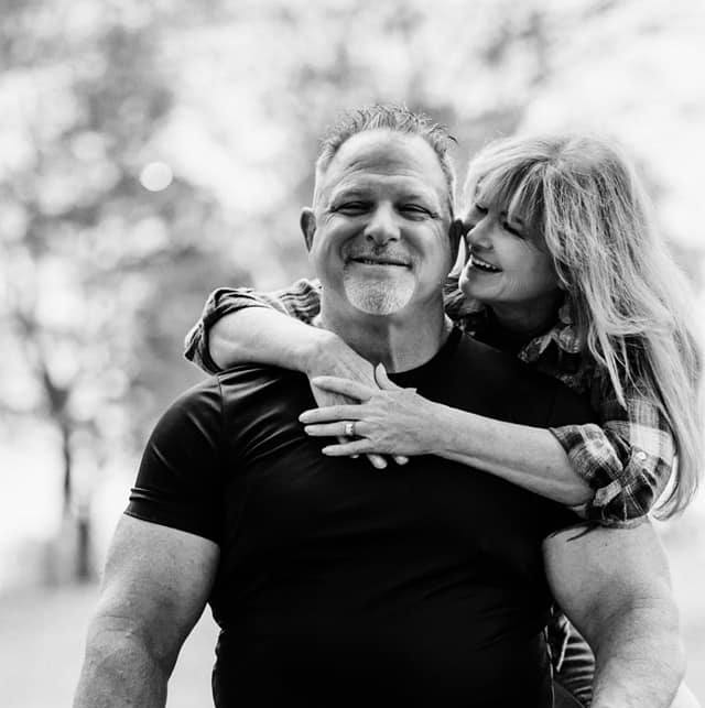 A man and a young girl are smiling and hugging outdoors, with trees in the background.