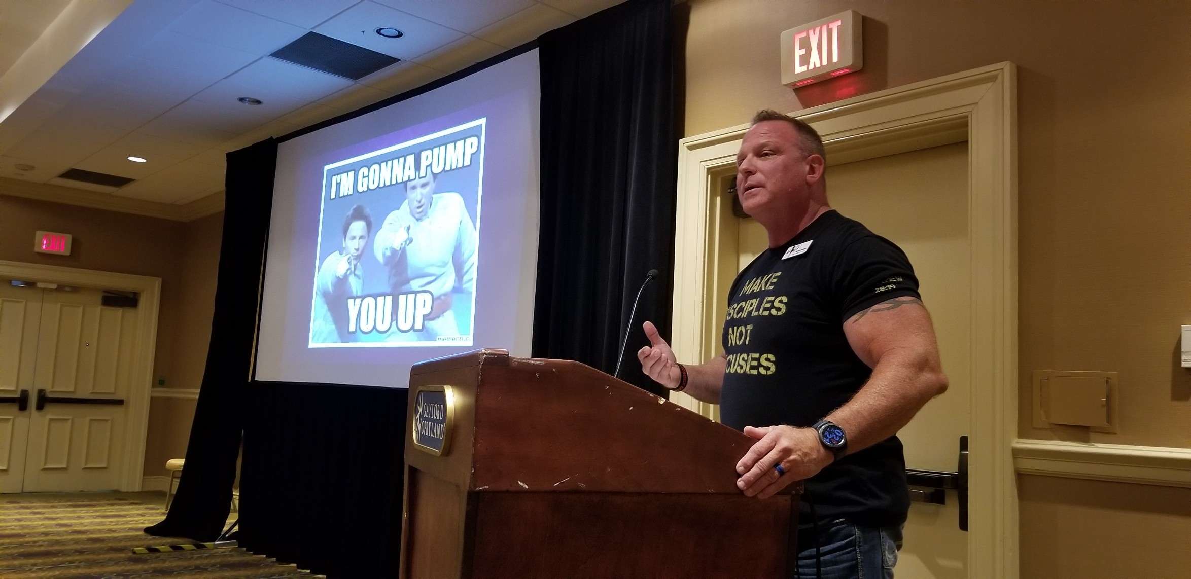 Man speaking at podium in a conference room with a presentation slide behind him, showing a humorous image and text.
