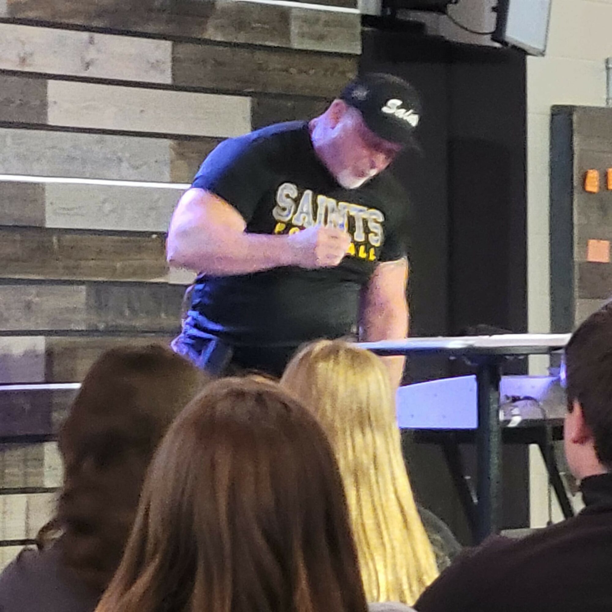 Person wearing a black cap and black t-shirt with yellow and white text, standing behind a table, in front of an audience.
