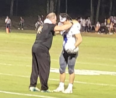 Person helping a young athlete adjust equipment on a sports field at night, with spectators in the background.