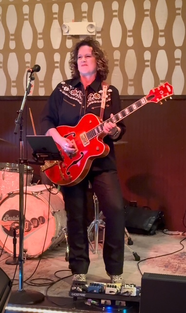 Person with curly hair playing a red electric guitar on stage, with patterned wallpaper background.