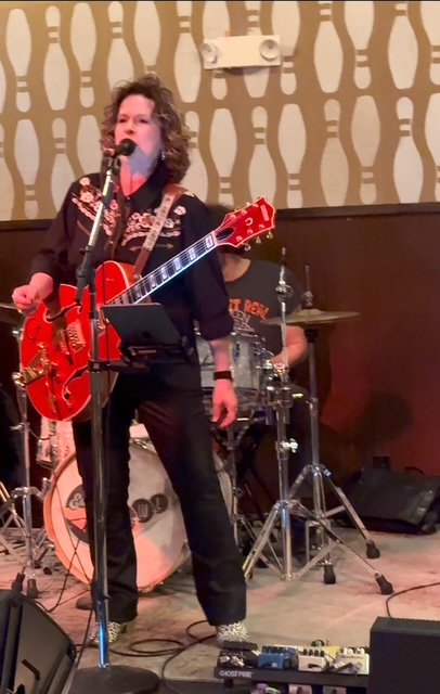 Person singing into microphone with guitar, band playing drums behind, patterned wallpaper background, performing on stage.