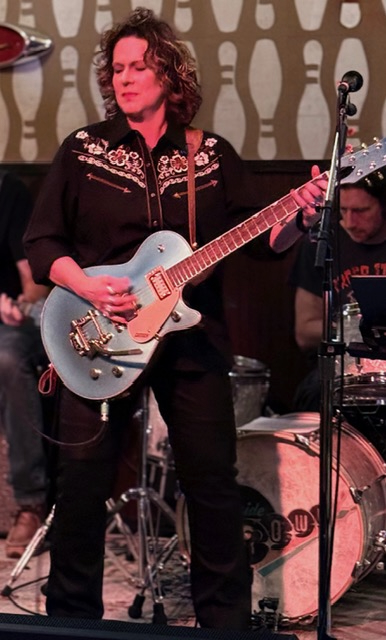 Woman with curly hair playing an electric guitar on stage, with a drummer in the background, patterned wall behind her.