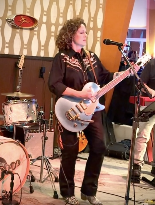 Woman with curly hair playing an electric guitar and singing into a microphone in a music venue.