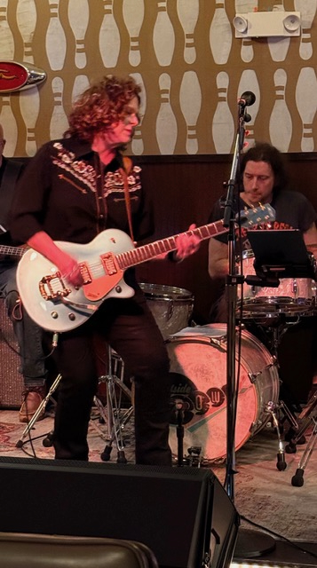 Two musicians perform on stage, one playing guitar and the other on drums, with patterned wallpaper in the background.