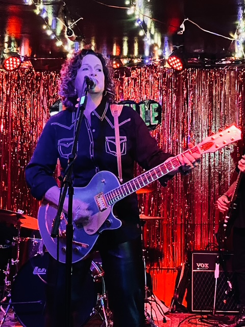 Woman singing and playing guitar on stage with red curtain background and stage lights.