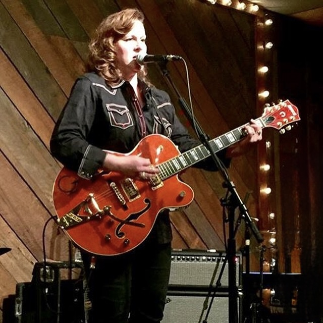 Woman playing an orange electric guitar and singing into a microphone on stage with wooden background and string lights.