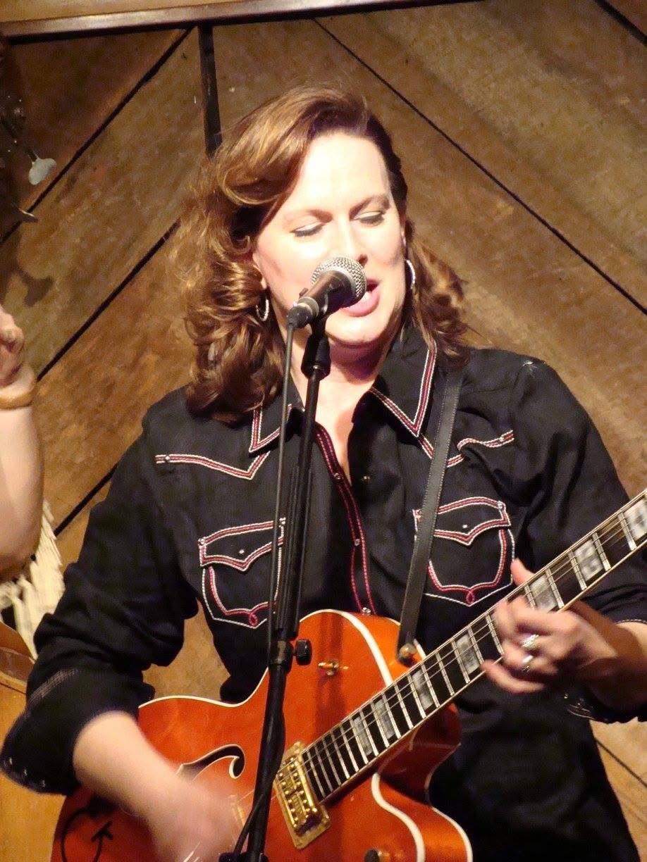 Woman singing into microphone while playing guitar on stage, with wooden background.