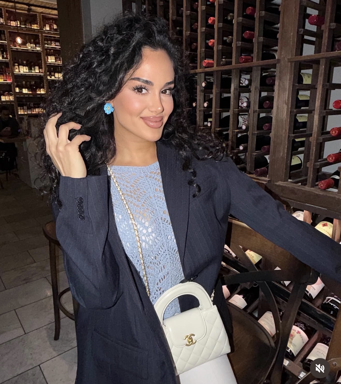 Woman with curly hair in a blazer and light blue top, holding a white handbag, in a wine cellar setting.