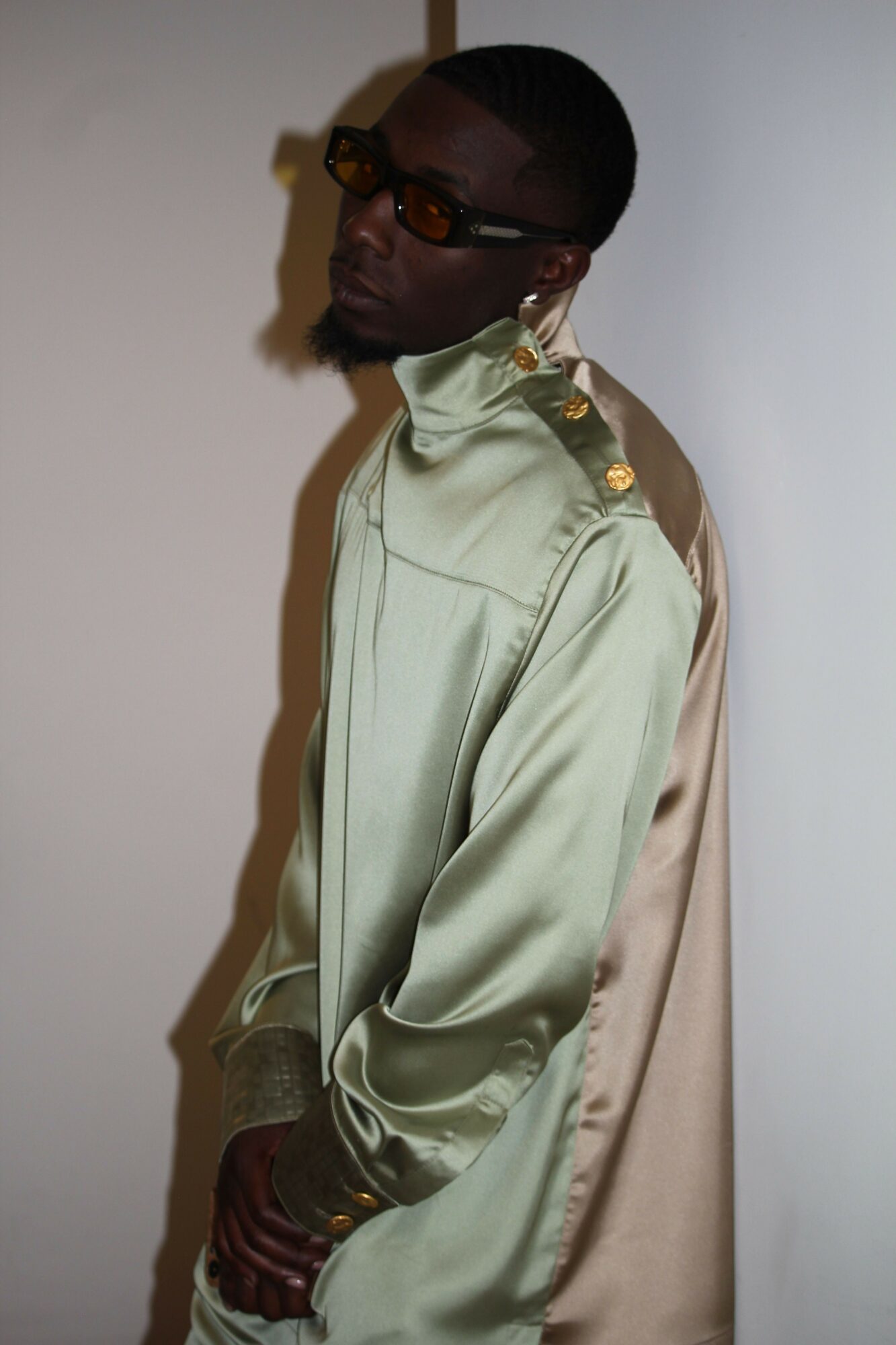 Person wearing sunglasses and a shiny jacket lying sideways on a surface, with a beige cloth behind them.