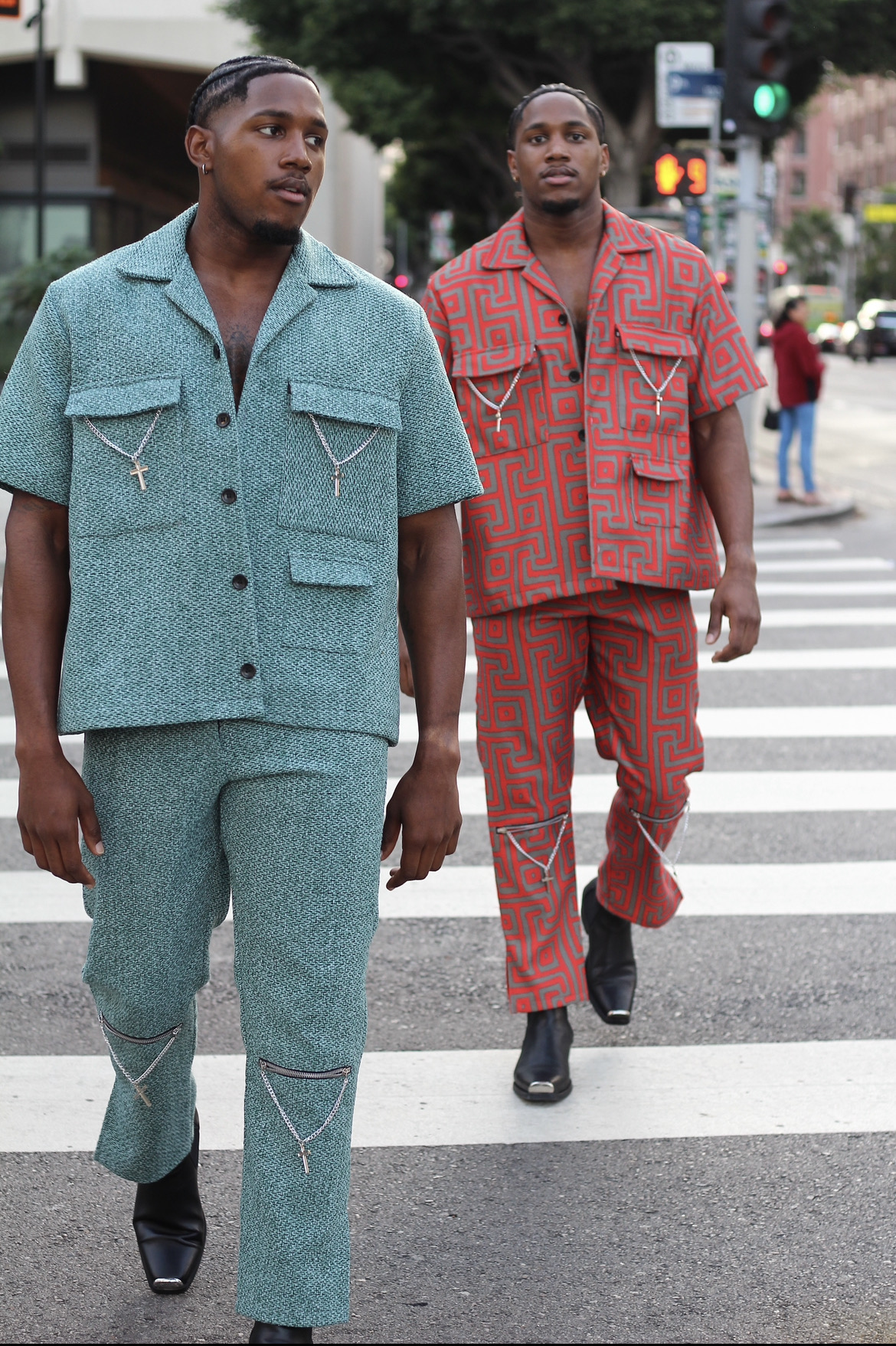 Two men walk across a crosswalk in an urban area, wearing colorful matching outfits, with buildings and traffic lights in the background.