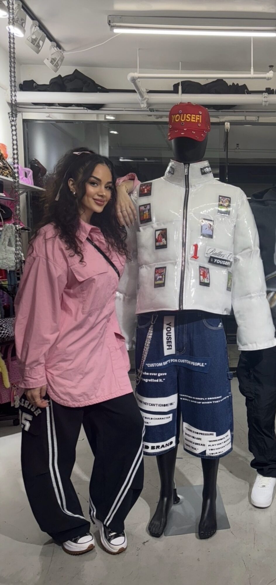 Woman in pink jacket and black pants standing next to a mannequin wearing a white jacket and blue shorts in a store.