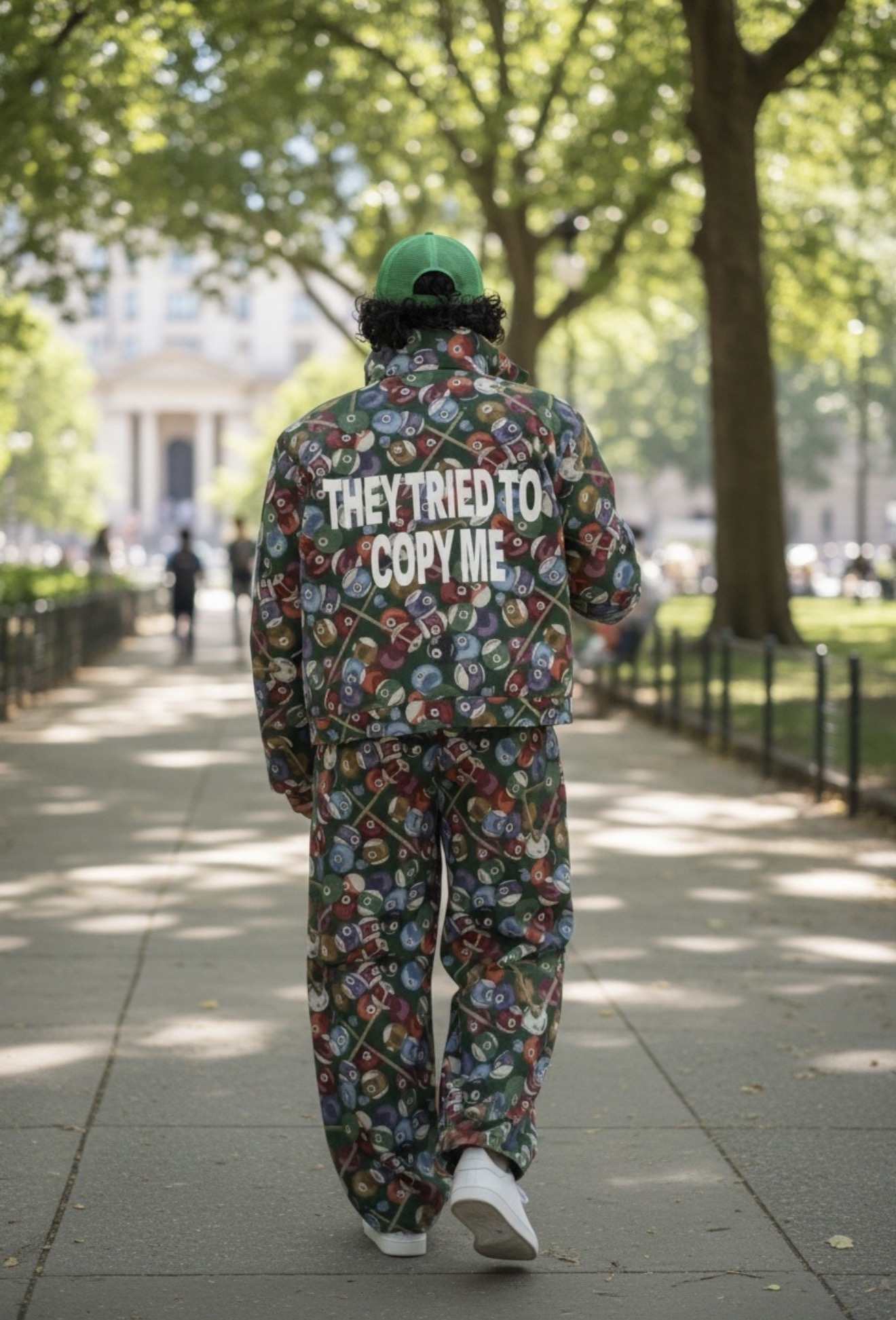 Person walking on a park sidewalk wearing a colorful patterned outfit and green cap, trees and buildings in background.