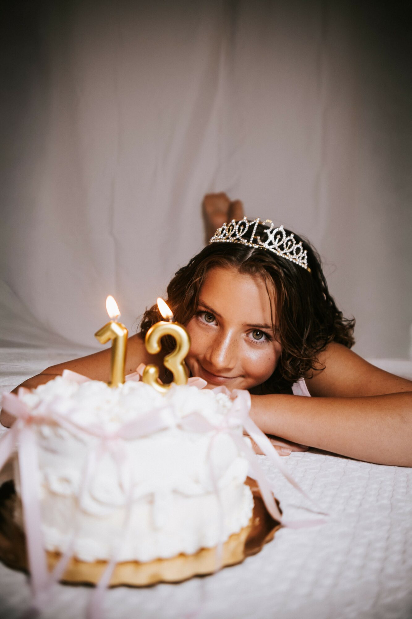 Young girl with a tiara resting her head on a table, birthday cake with candles in front, white background.
