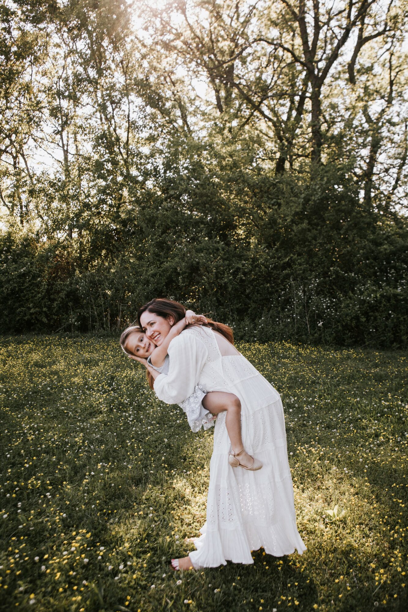 Two women in white dresses outdoors, one lifting the other, smiling, with trees and grass in background.