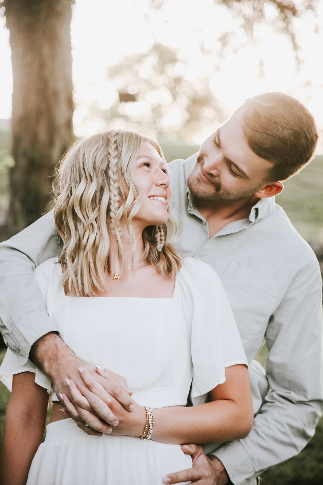 A man and woman outdoors, smiling and looking at each other, with trees and sunlight in the background.