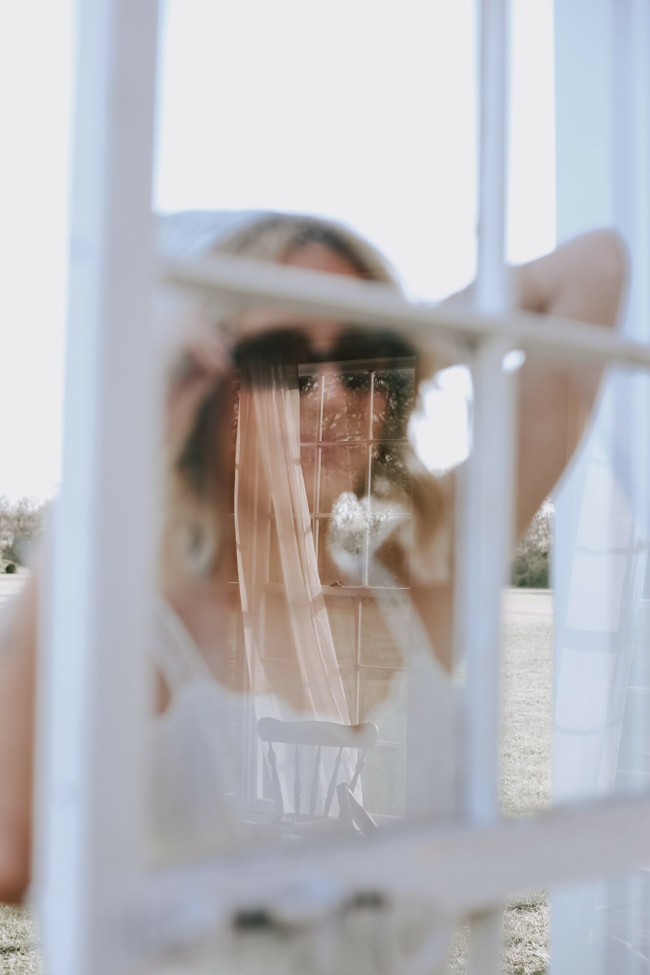 A woman with a veil looks through a window with white frames, her face partially obscured by the glass and curtains.