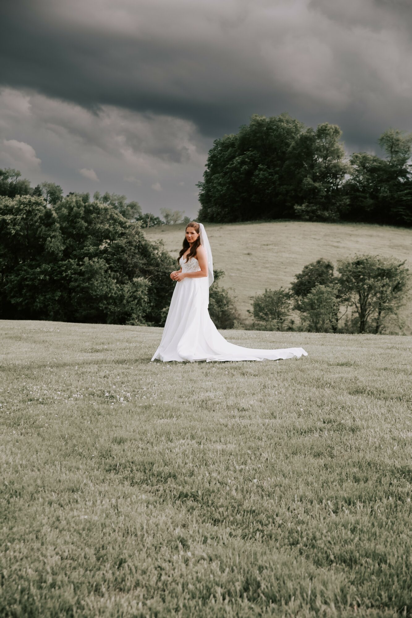 A woman in a white wedding dress stands on a grassy field with trees and dark clouds in the background.