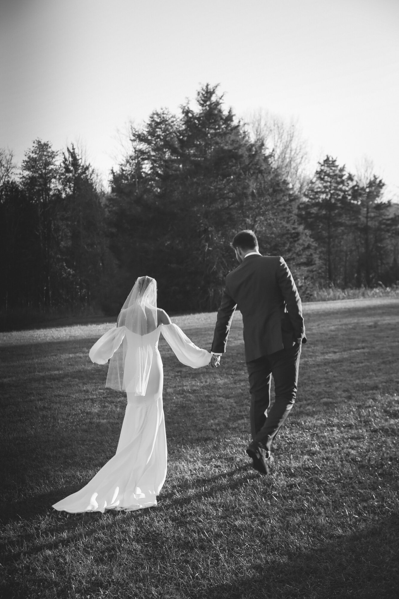 A couple holding hands walking outdoors, woman in a long dress and veil, man in a suit, trees in background.