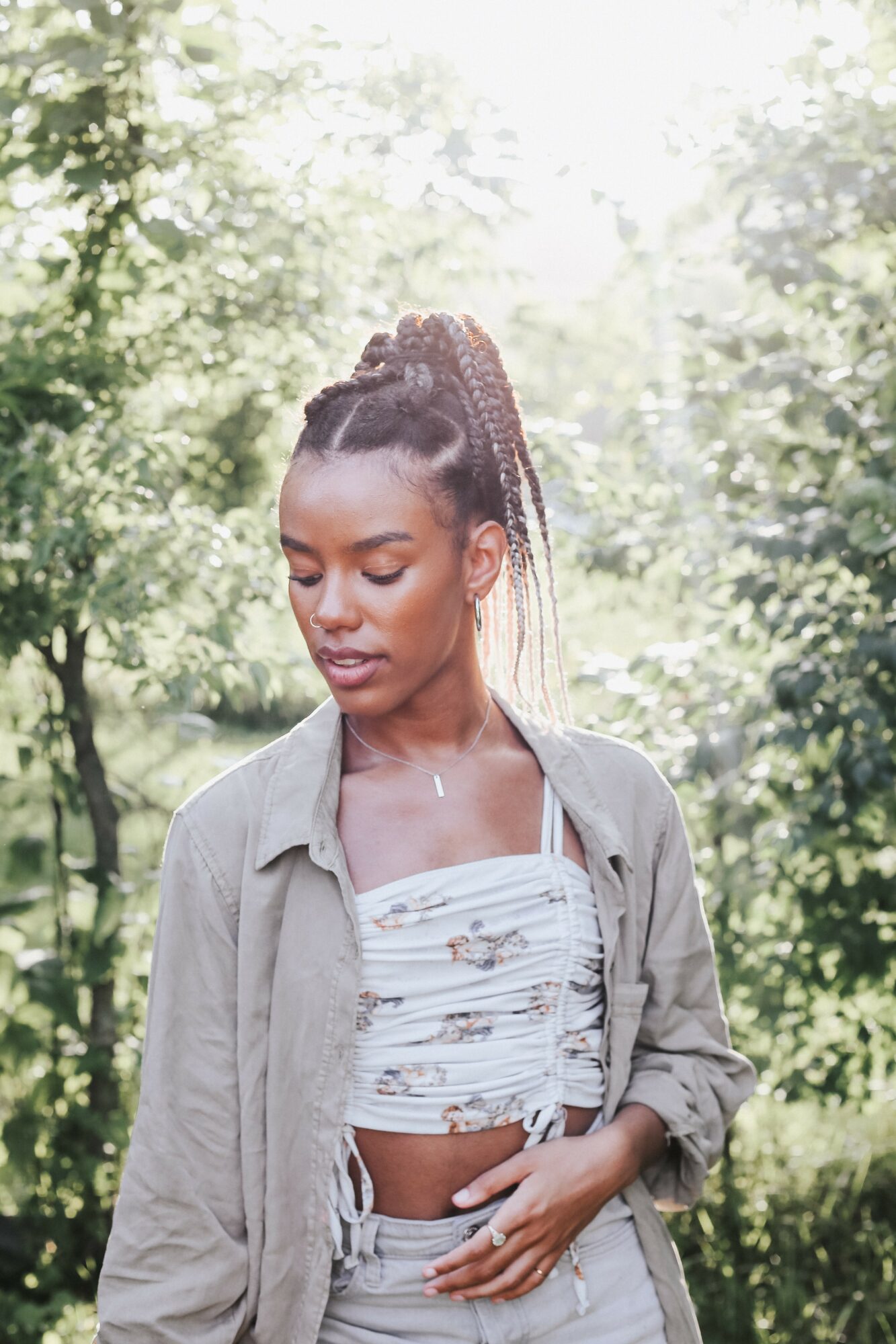 Young woman with braided hair standing outdoors among trees, wearing a light jacket and patterned crop top.